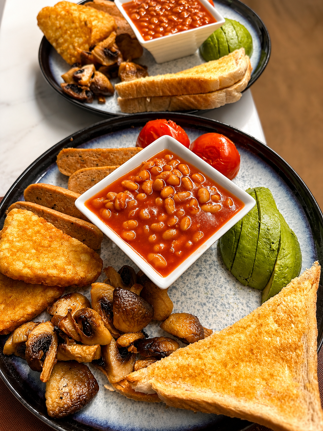 Two full breakfast plates with beans, avocado, mushrooms and toast